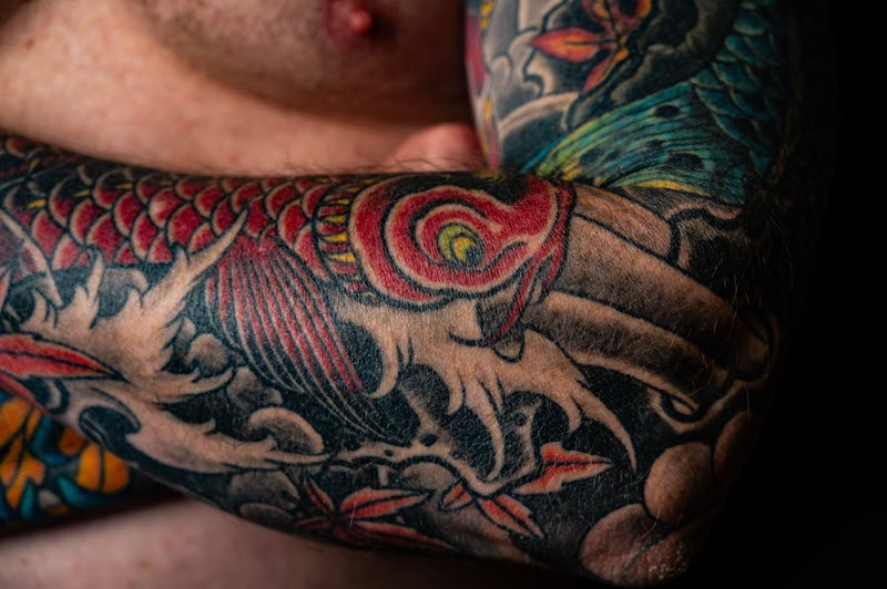 Close-up of a red koi fish tattoo on a man's arm. The tattoo is detailed and colorful, and it covers most of the man's forearm.