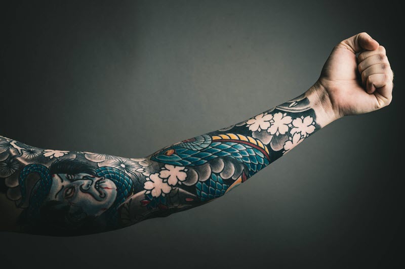 A man's arm with a full sleeve tattoo of a dragon and flowers. The dragon is green and red, with scales and wings. 