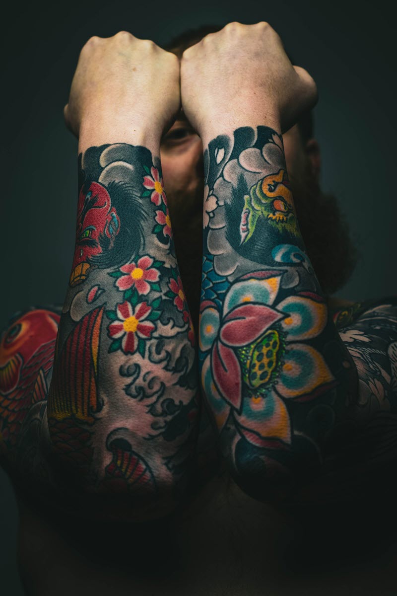  A man with a lot of tattoos on his arms is looking at the camera. The tattoos are colorful and intricate, and they cover most of his arms.