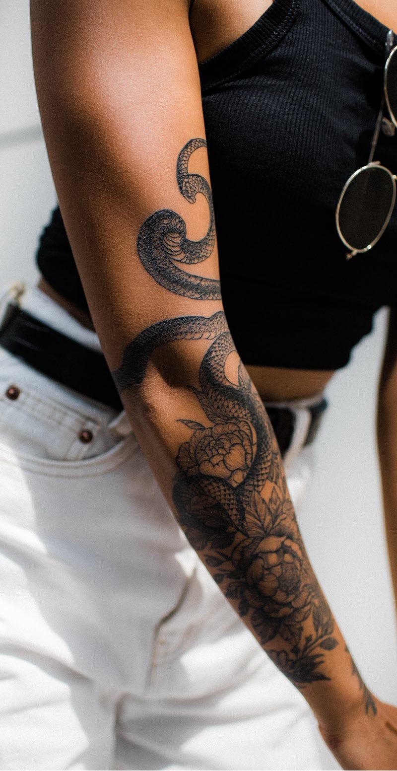 A woman has a snake tattoo on her right arm, which is coiled around her arm. 